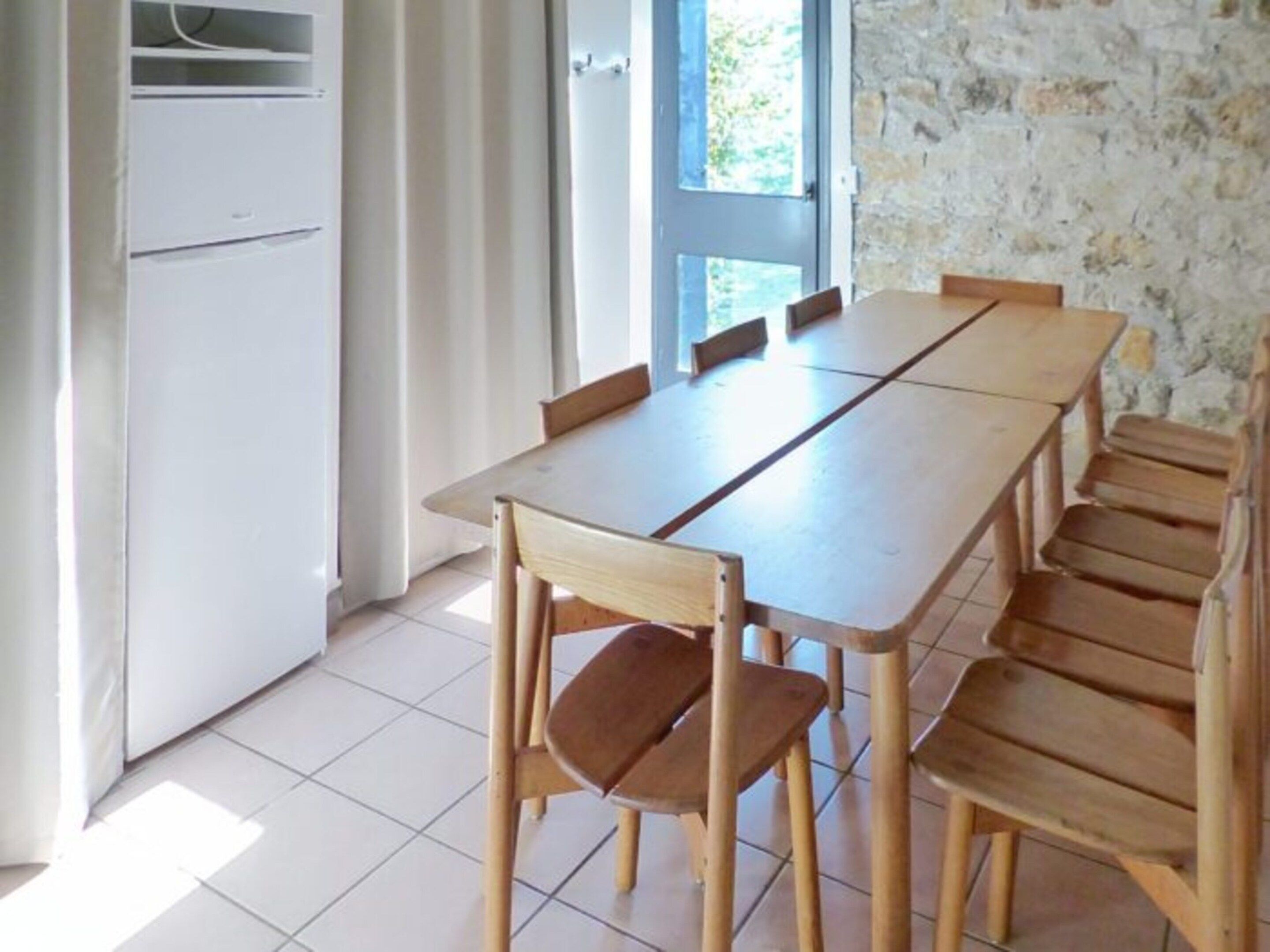 Un coin repas avec une longue table et des chaises en bois, un réfrigérateur blanc et un mur en pierre.