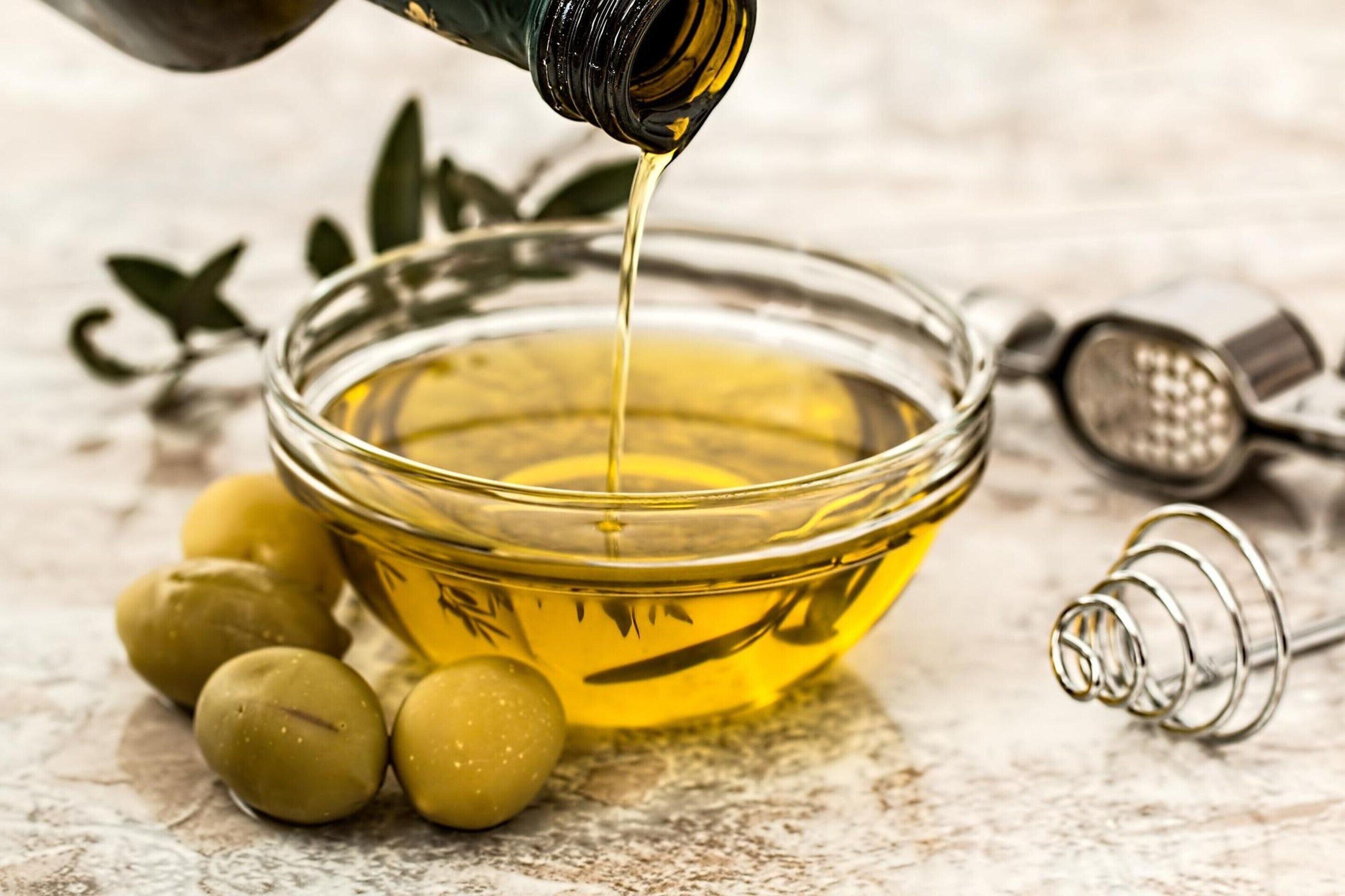 Un cuenco de vidrio con aceite de oliva siendo vertido, rodeado de aceitunas y una rejilla metálica.