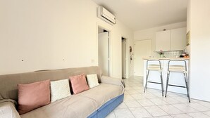 A living room with a sofa, two chairs, a kitchen area with a counter and cabinets, and an air conditioning unit on the wall.