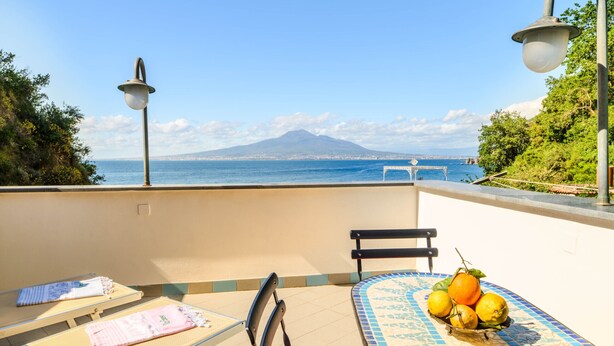 Un balcone con vista su una montagna e il mare, un tavolo in maiolica con una ciotola di frutta e due lettini prendisole.