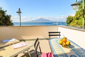En balkon med udsigt til et bjerg og havet, et flisegulvsbord med en skål med frugt, og to liggestole.