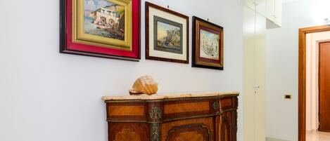 A hallway with framed artwork on the wall, a wooden cabinet with a decorative shell, and a door in the background.