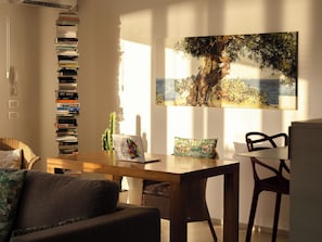 A cozy living room with a bookshelf, a cactus, a laptop, a dining table, and a painting of a tree by a sea.