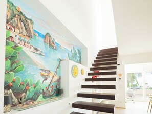 A staircase with dark steps leading to a landing, a mural with cacti and boats, and a glimpse of a balcony with outdoor furniture.