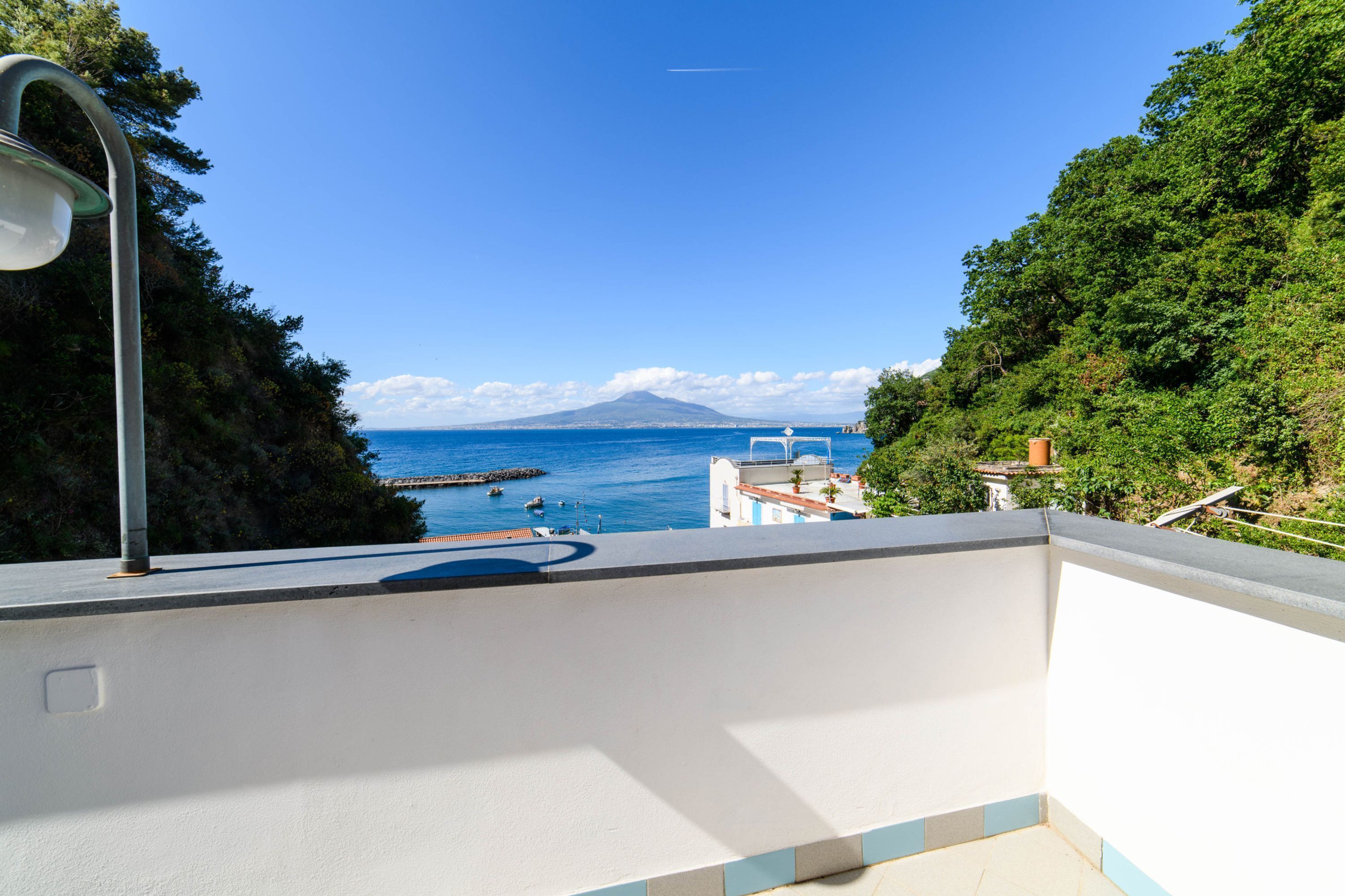 A balcony with a view of the sea, a mountain in the distance, and lush greenery.