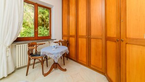 Quarto com guarda-roupa de madeira, uma mesa com toalha rendada, duas cadeiras, janela com vista para vegetação e radiador.
