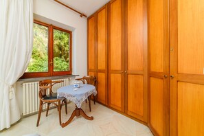 Een kamer met een houten garderobe, een tafel met kantkleed, twee stoelen, een raam met uitzicht op groen, en een radiator.