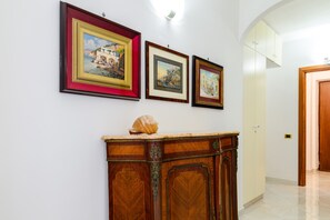 Een gang met ingelijste kunstwerken aan de muur, een houten kastje met een decoratieve schelp, en een deur naar een andere kamer.
