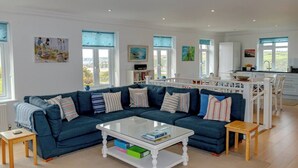 A spacious living room with a large sectional sofa, a glass coffee table, and a kitchen area with white cabinets and a dining table. - 11 The Downs, a coastal retreat in Thurlestone South Devon (Thurlestone)