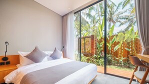 Um quarto moderno com uma cama grande, uma mesa de cabeceira, uma escrivaninha e vista para um jardim tropical.