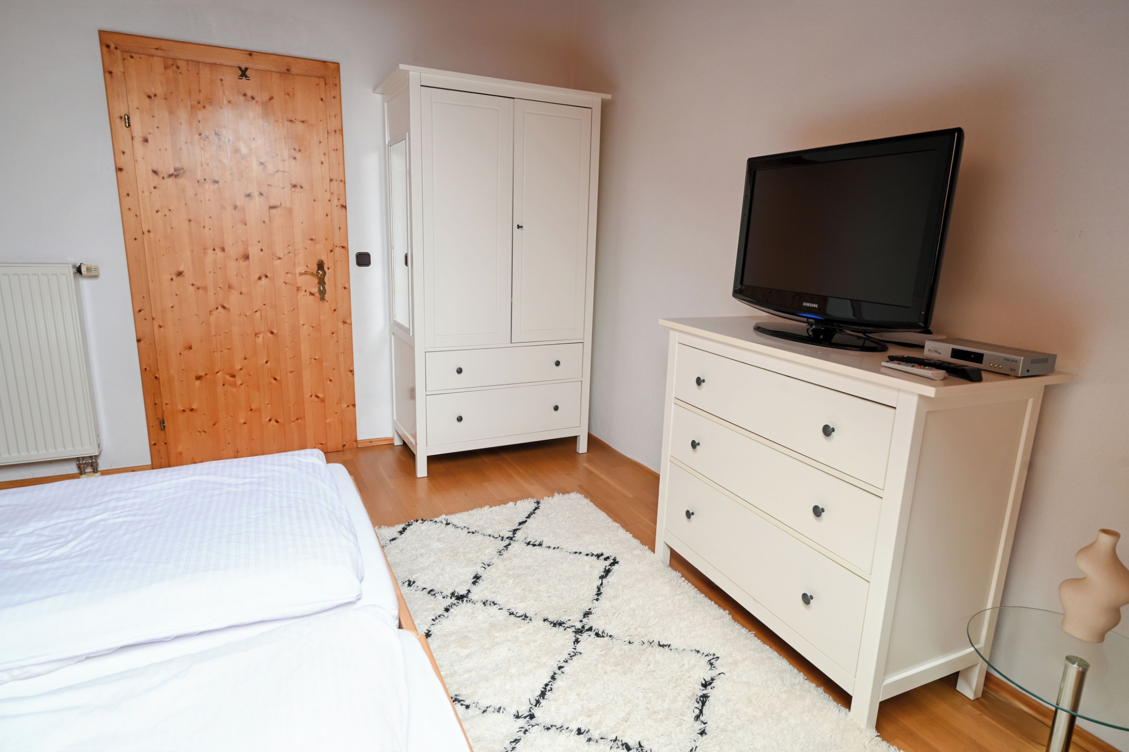 A bedroom with a bed, a TV, a white wardrobe, and a wooden door.