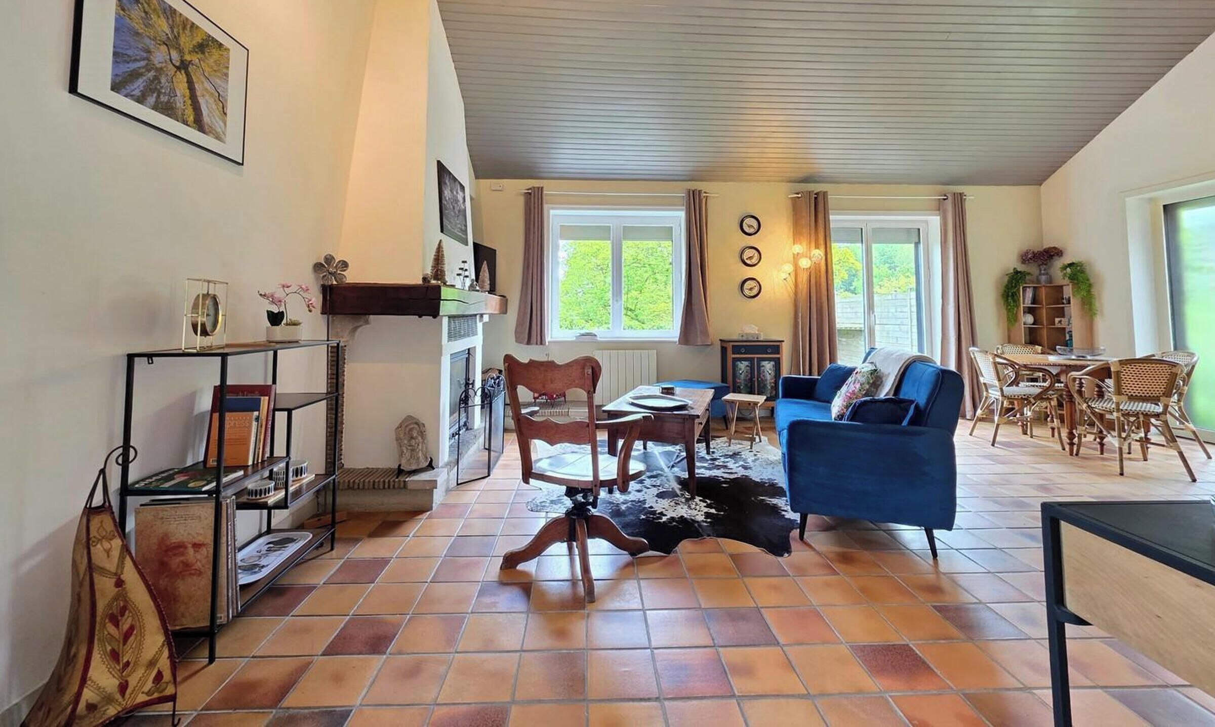 Una sala de estar con un sofá azul, sillas de madera, una mesa de centro, una chimenea y un ventanal con cortinas.