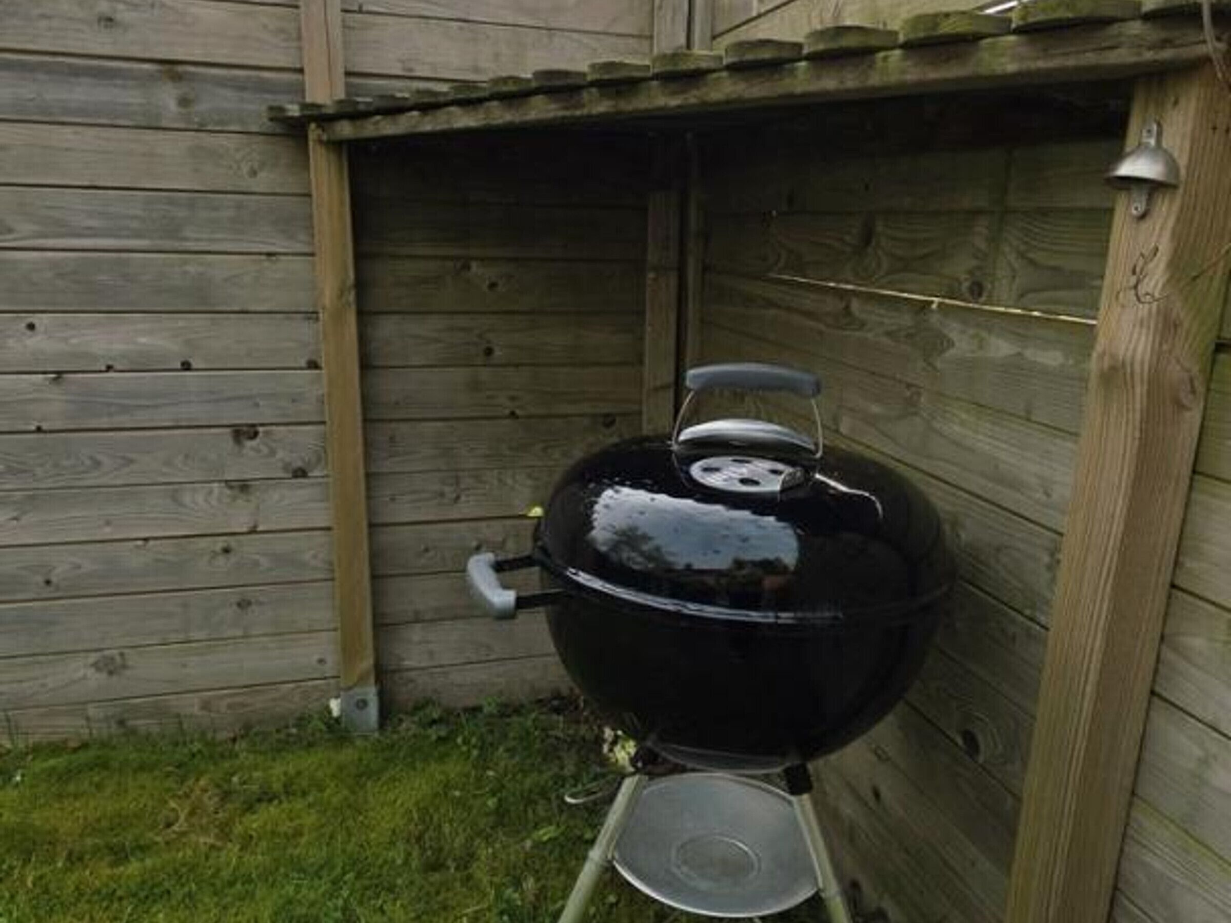 A black outdoor grill with a metal bowl underneath, positioned against a wooden fence.