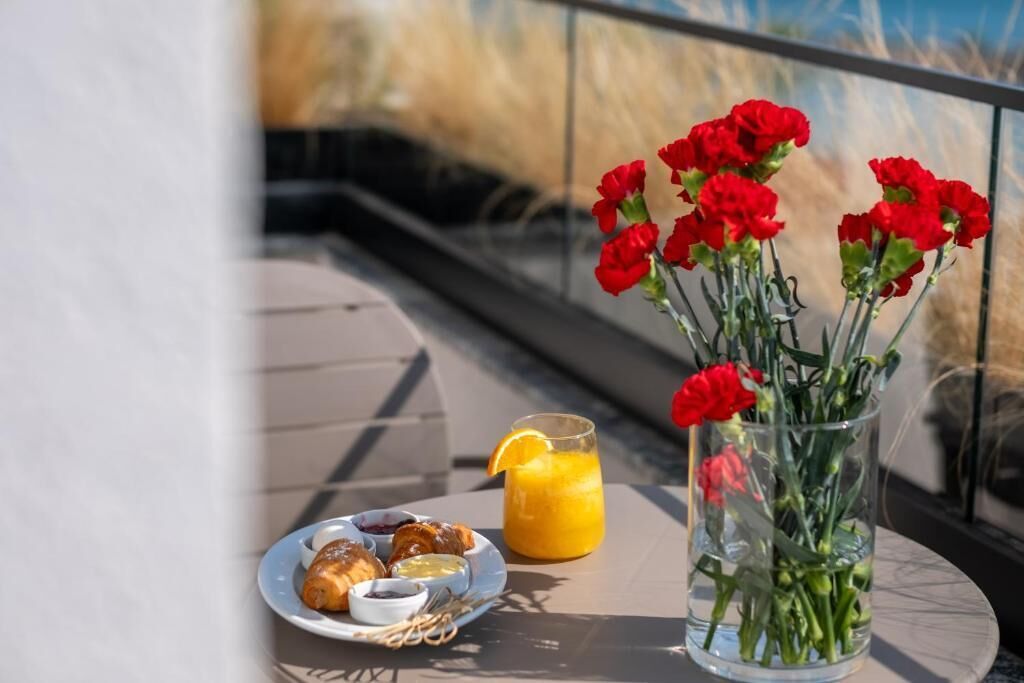 Una mesa con un plato de pasteles, un vaso de zumo de naranja y un jarrón con flores rojas.
