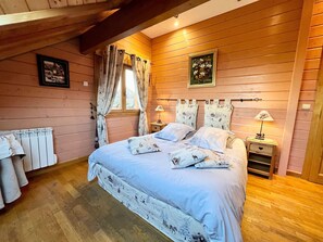 A bedroom with a wooden ceiling, a bed with floral bedding, a window with curtains, a bedside table with a lamp, and a radiator.