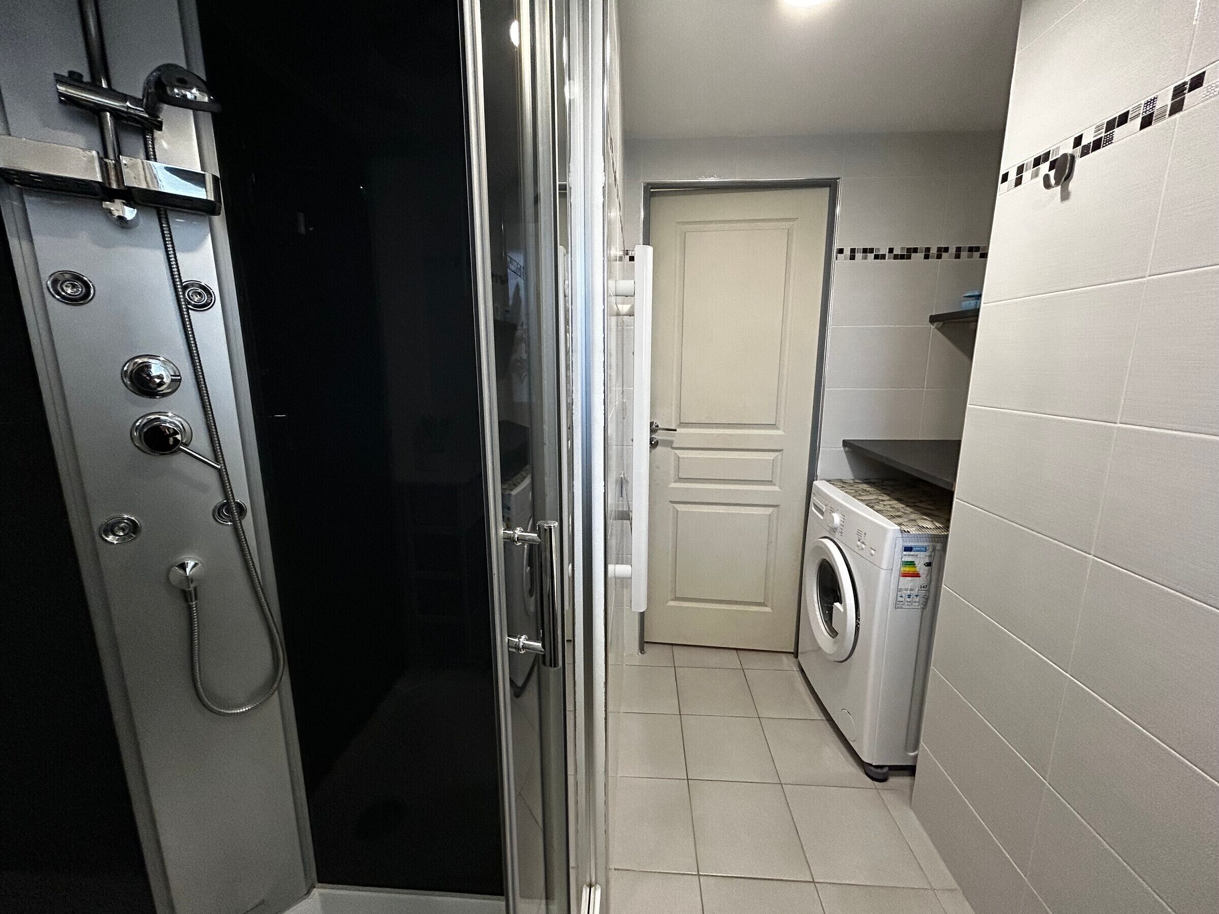 A compact bathroom with a glass-enclosed shower, a washing machine, and a door leading to another room.