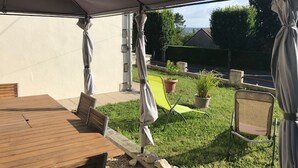 Un patio al aire libre con una mesa y sillas de madera, un área cubierta con un toldo y un jardín césped con plantas en macetas.