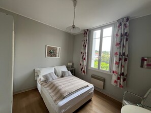 Ein Schlafzimmer mit Bett, Nachttisch, Stuhl, einem Fenster mit Vorhängen, Heizkörper und Deckenventilator.