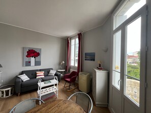 Ein modernes Wohnzimmer mit einer Couch, einem Esstisch und einem Schaukelstuhl. Ein Heizkörper, ein kleiner Beistelltisch und ein Fenster mit Vorhängen sind ebenfalls vorhanden.