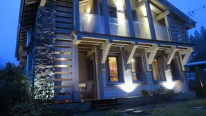 Una cabaña de madera de dos pisos con fachada de piedra, ventanas grandes y un porche con mobiliario exterior.
