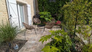 Um pátio de pedra com cadeiras de vime, uma mesa pequena e um jardim com diversas plantas e uma cerca de arame.