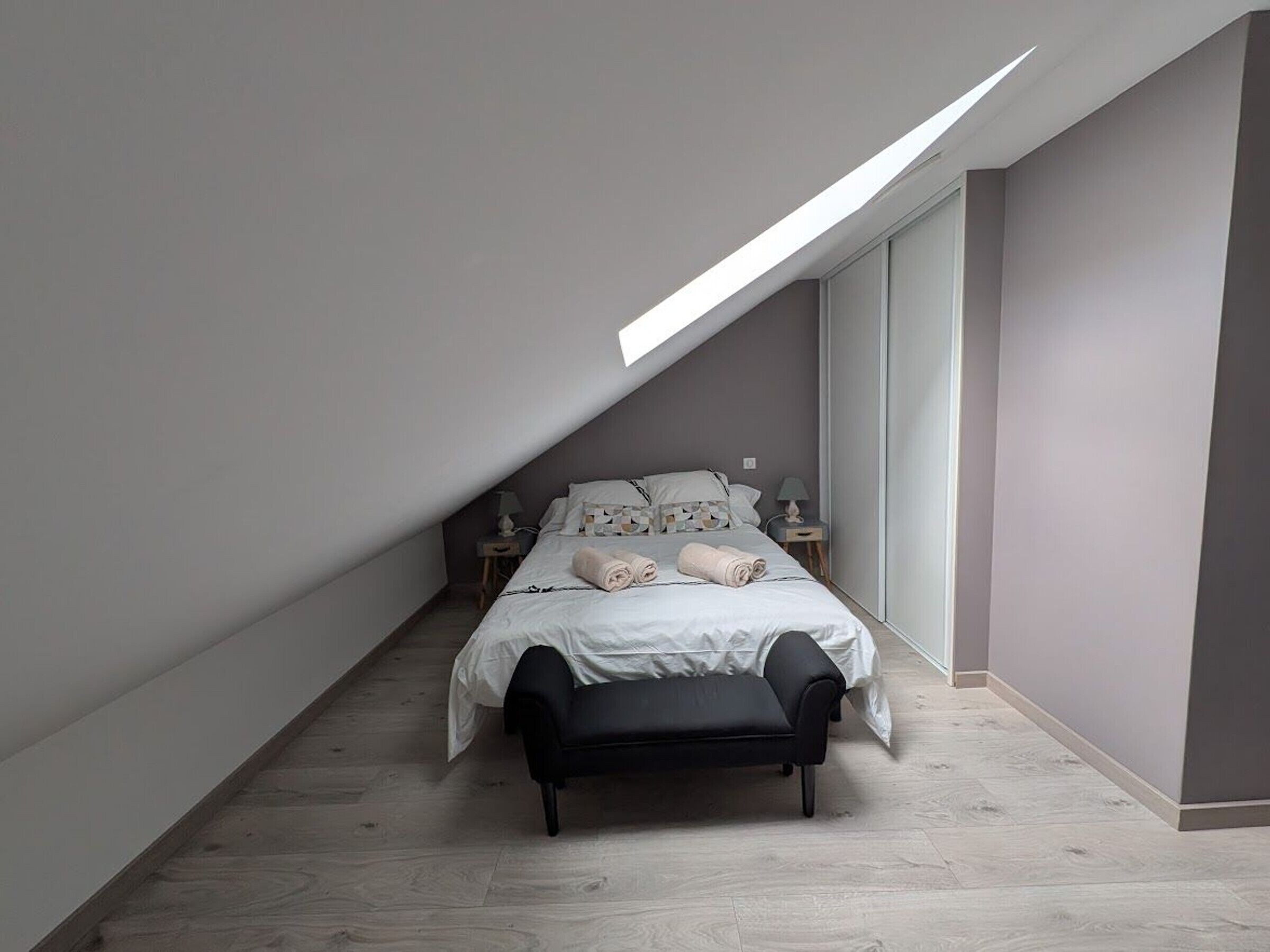 Une chambre avec un plafond en pente, un lit avec une literie blanche, un banc noir et une table de chevet avec une lampe.