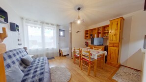 A cozy living room with a sofa, dining table, chairs, and a kitchen area with wooden cabinets and a television. - Quiet apartment, private parking - 4 guests (Les Rousses)