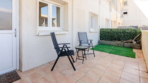 Eine Terrasse mit zwei Stühlen, einem kleinen Tisch, einem Fenster und einer grünen Hecke.