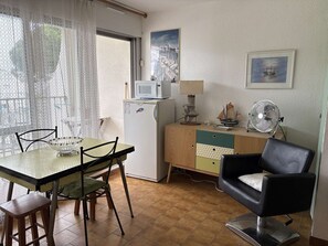 A compact room with a dining table, chairs, a small refrigerator, a microwave, a cabinet, a fan, and two wall-mounted pictures. - Apartment with canal view - 4 guests (Port Camargue)