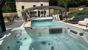 A hot tub with a glass enclosure, a pool, and lounge chairs in a backyard setting.