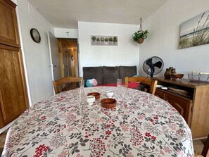 A dining area with a floral tablecloth, wooden chairs, a clock, a cross on the wall, and a fan. - Apartment with terrace and parking - 4 guests (Cap d'Agde)