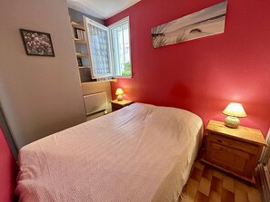 Ein Schlafzimmer mit Bett, Nachttisch mit Lampe, einem Fenster mit Vorhängen, einem Bücherregal und einer Heizung.