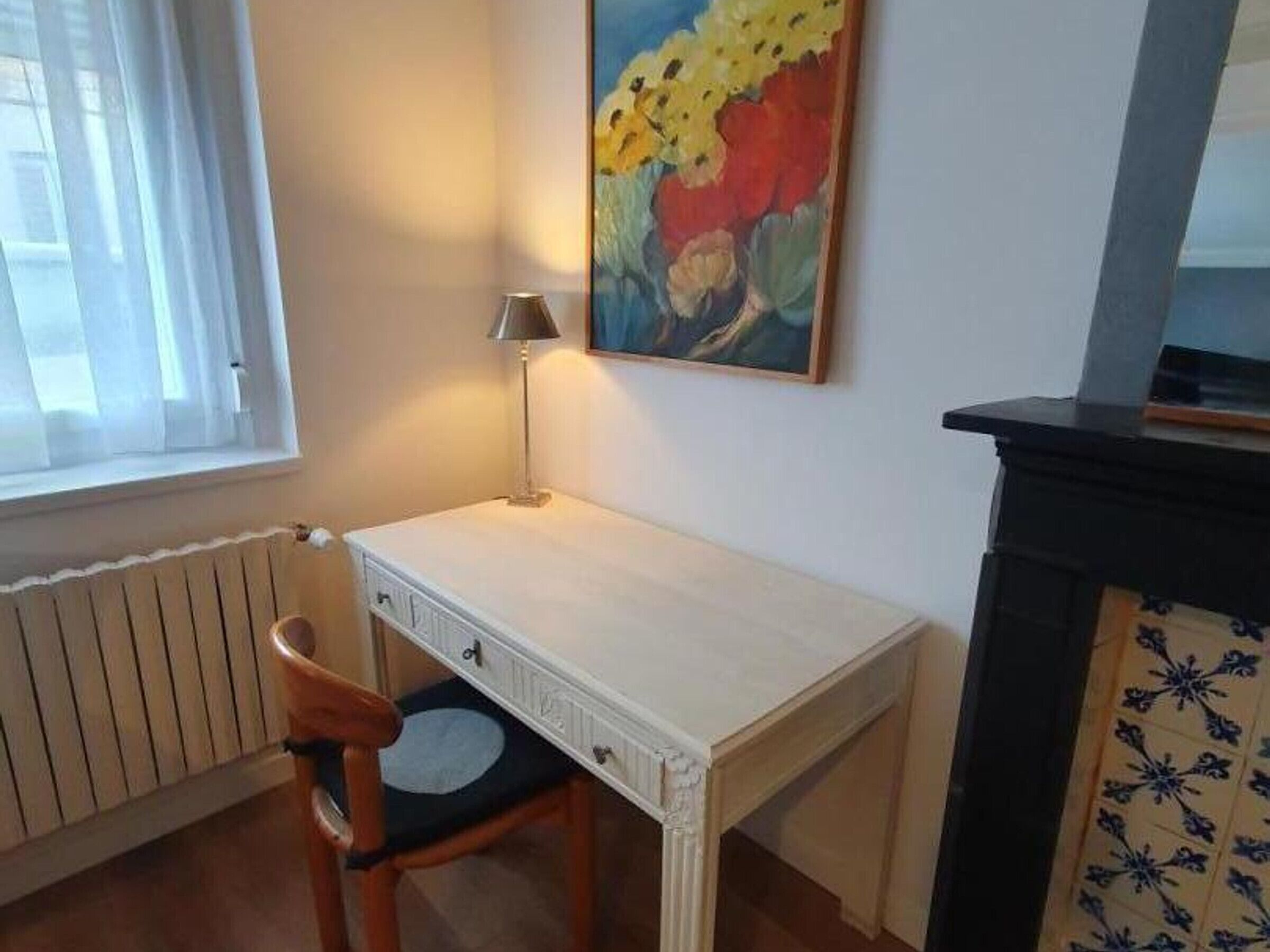 Une pièce avec un bureau blanc, une chaise, une lampe, un tableau, un radiateur et une cheminée recouverte de carreaux.