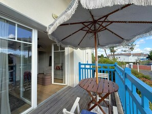 Eine Terrasse mit Tisch und Sonnenschirm, ein blauer Zaun und eine Glastür, die zu einem Innenraum führt.