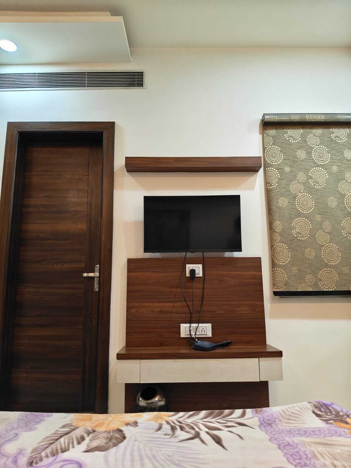 A bedroom with a wooden door, a TV mounted on a wooden shelf, a power outlet, and a patterned curtain.