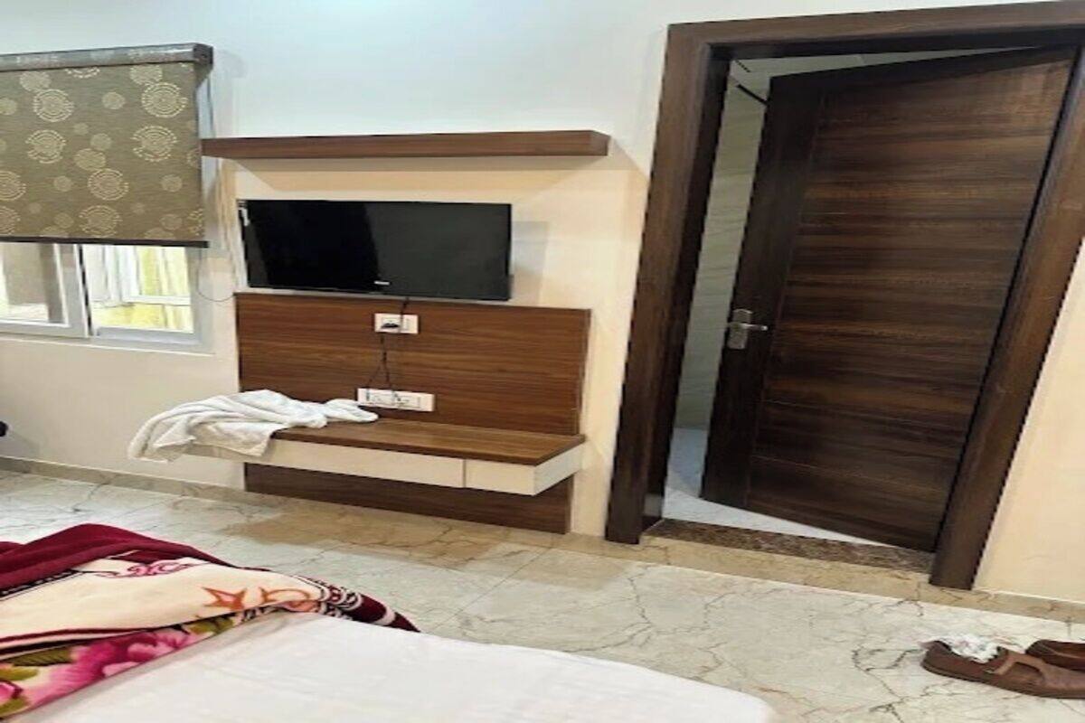 A bedroom with a bed, a TV mounted on a wooden shelf, a door, and a window with curtains.