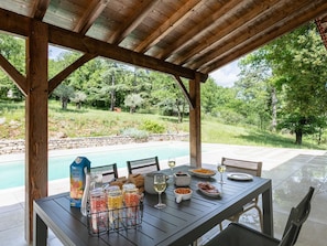 A covered outdoor dining area with a pool view, wooden beams, and a stone wall.
