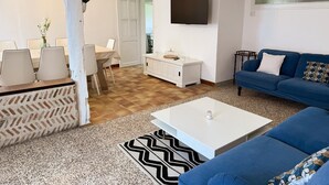 Un salon moderne avec un canapé bleu, une table basse blanche, un téléviseur fixé au mur et un coin repas comprenant une table et des chaises.