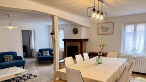 Un espace repas avec une longue table, des chaises blanches, un lustre et une cheminée.