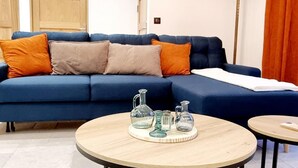 Un canapé bleu agrémenté de coussins orange et beige, une table basse ronde en bois surmontée de verrerie, et une petite table ronde recouverte d’une nappe blanche.