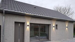 Un bâtiment moderne avec un toit recouvert de tuiles grises, de grandes portes vitrées et deux lampes fixées aux murs.