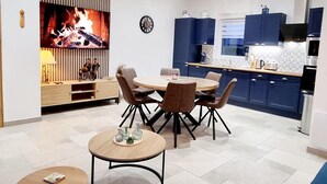 Une cuisine moderne avec des meubles bleus, un coin repas avec une table ronde et des chaises, un téléviseur fixé au mur et une horloge au mur.