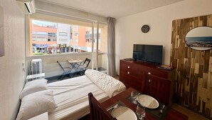 A hotel room with a bed, a TV, a dining table with plates and glasses, a wooden wall panel, and a window with a view of buildings. - Studio in Cap d'Agde center - 2 guests (Cap d'Agde)
