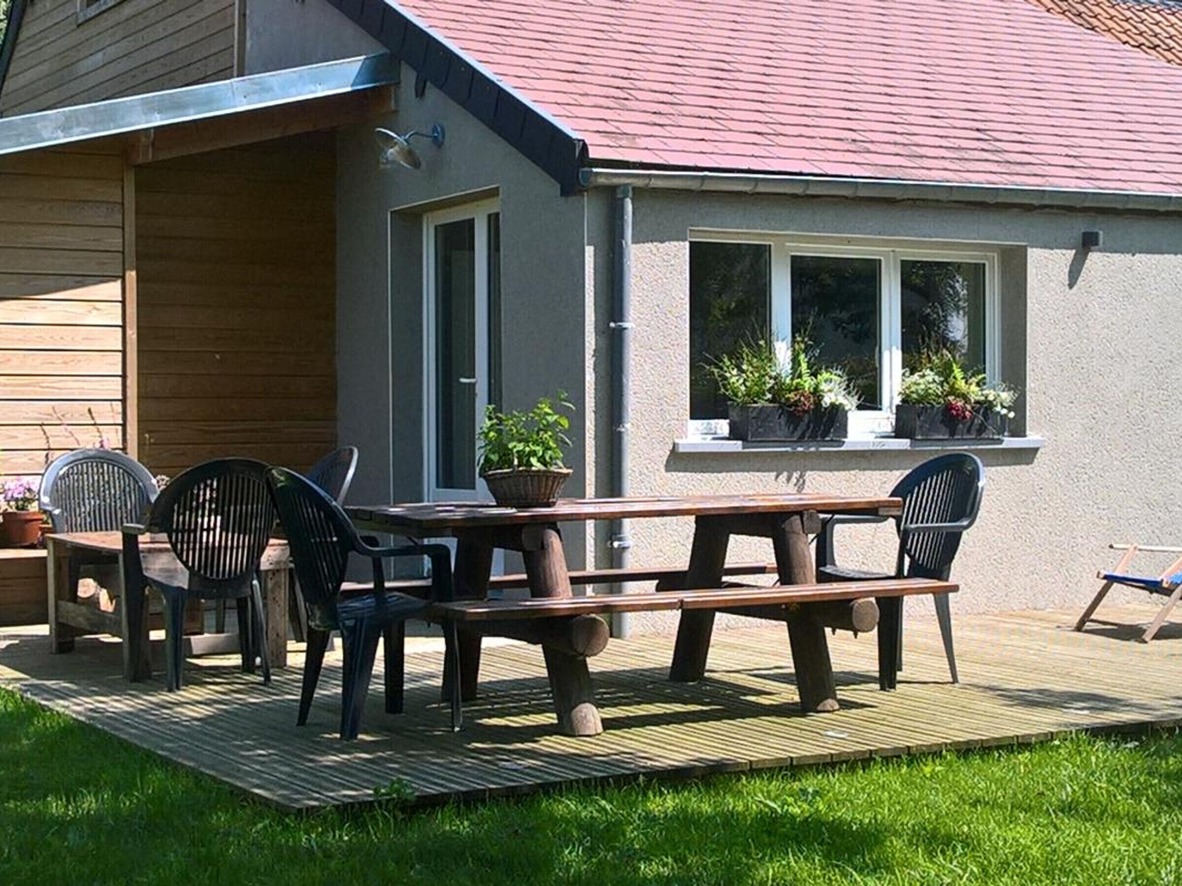 A wooden deck with a picnic table and chairs, potted plants, and a building with a red roof.