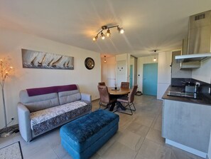 Ein modernes Wohnzimmer mit einer Couch, Hocker, Esstisch und Stühlen. Ein Küchenbereich ist erkennbar, mit einem Herd und Schränken.