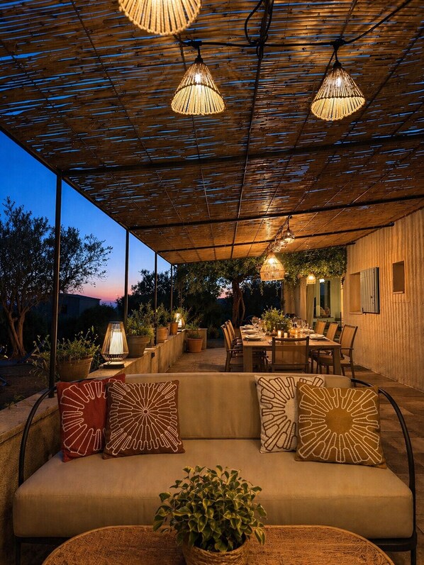 Eine Terrasse im Freien mit einer Holz-Pergola, hängenden Lichtern, einem Esstisch für vier Personen und einer gepolsterten Sitzecke mit gemusterten Kissen.