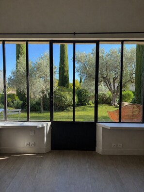 Ein Zimmer mit großen Fenstern, Holzboden und Blick auf einen Garten mit Bäumen und einer Bank.