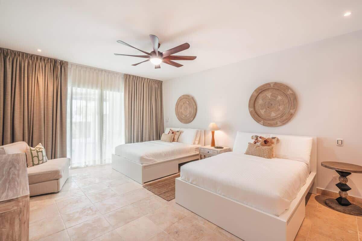 A hotel room with two beds, a ceiling fan, a large window with curtains, a bedside table with a lamp, and a round table.