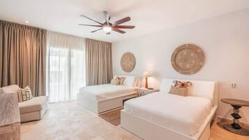 A hotel room with two beds, a ceiling fan, a large window with curtains, a bedside table with a lamp, and a round table.