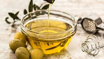 Una ciotola di vetro trasparente con un liquido giallo, probabilmente olio, olive e un contenitore metallico sullo sfondo.
